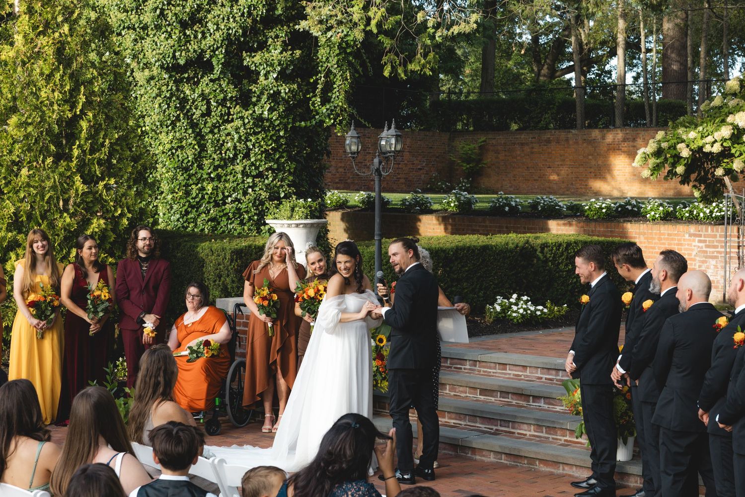 Outdoor wedding ceremony with bride and groom at altar, bridesmaids in yellow and orange dresses, groomsmen in black suits.