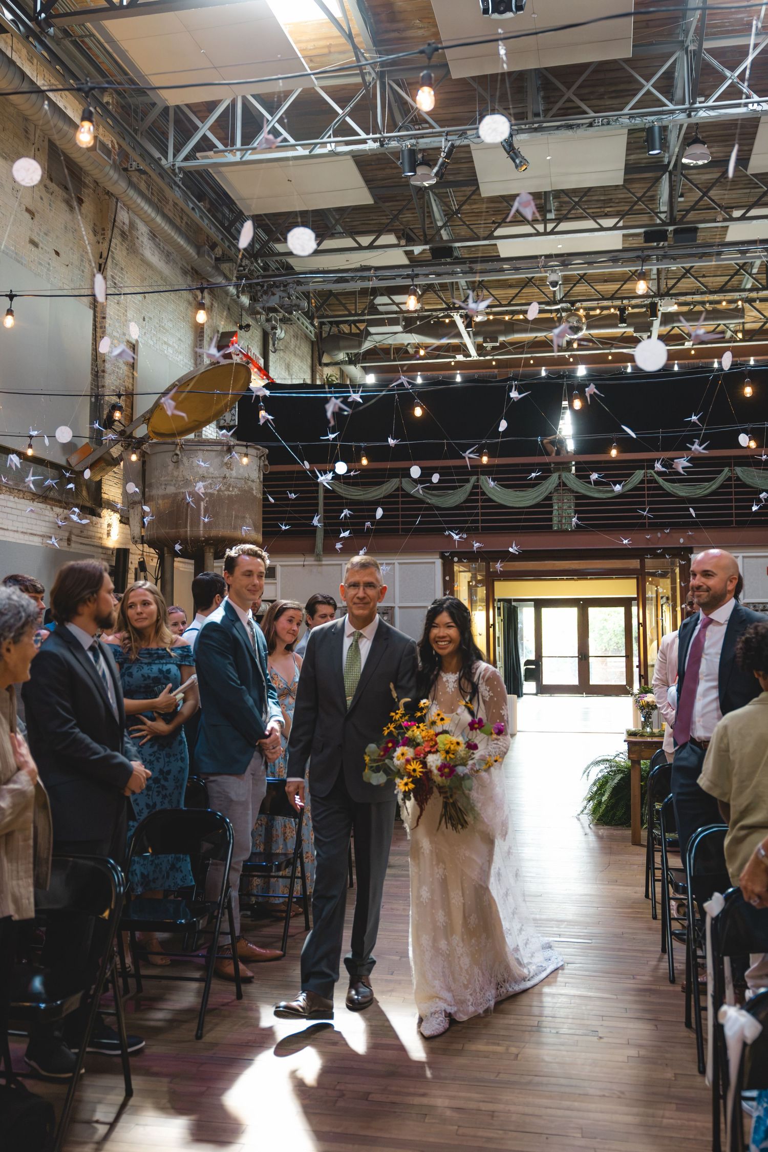 Bride walks down the aisle with her father in a rustic industrial wedding venue decorated with string lights.