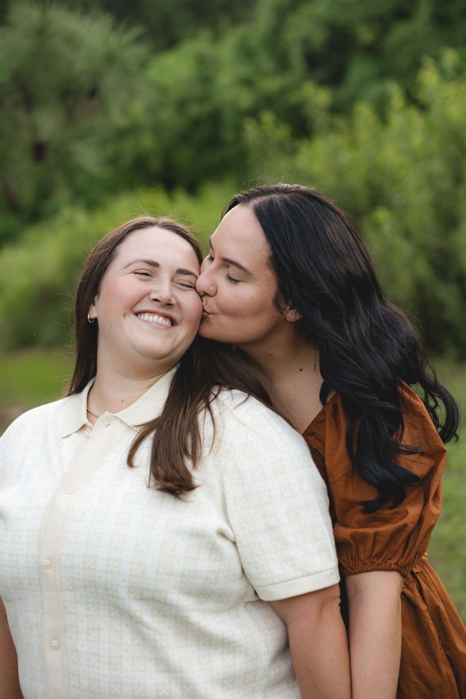 Two women share a tender kiss outdoors, one smiling in white dress, other in rust dress, green trees behind.