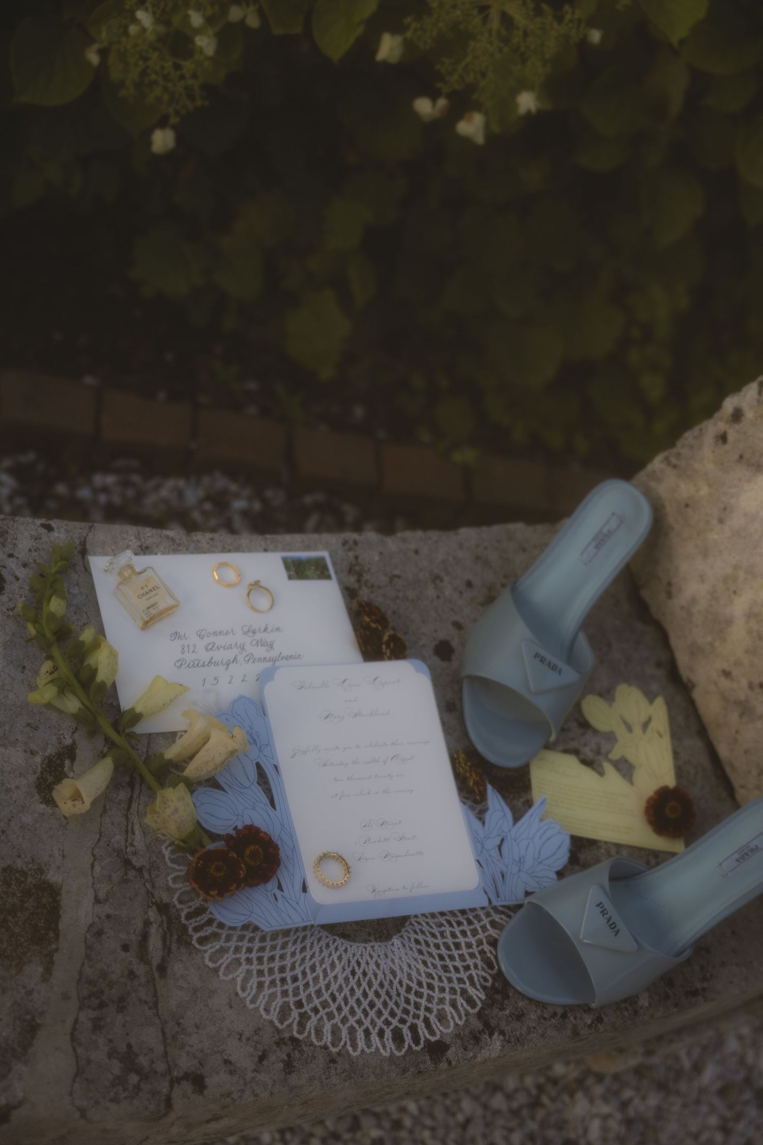 Wedding flat lay with blue sandals, invitation suite, gold rings, and floral accents on stone steps.