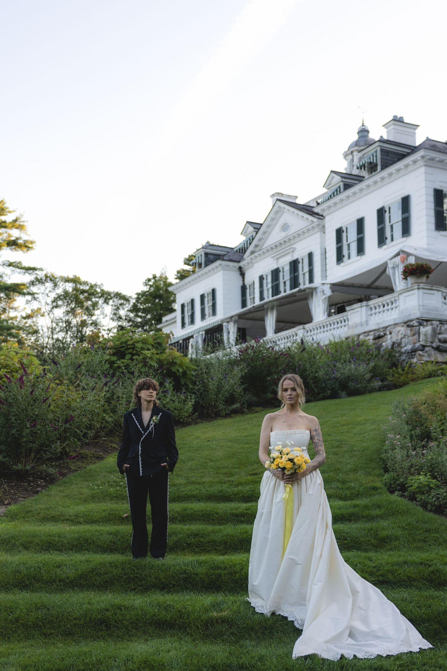 Two brides pose on green lawn steps before a white colonial mansion, one in a suit, one in a white gown with yellow bouquet.