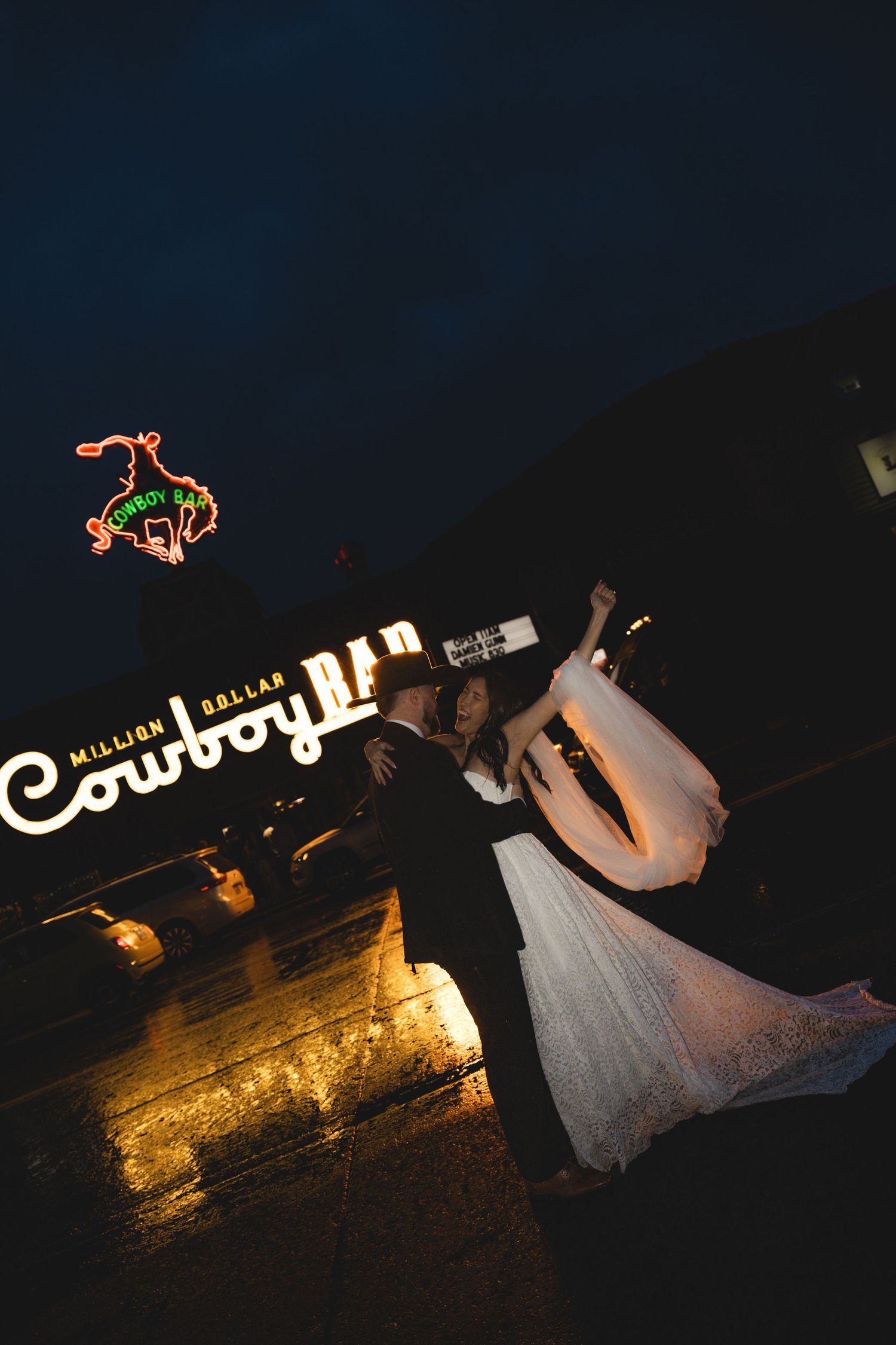 Bride and groom embracing outside the Million Dollar Cowboy Bar at night with neon signs glowing.