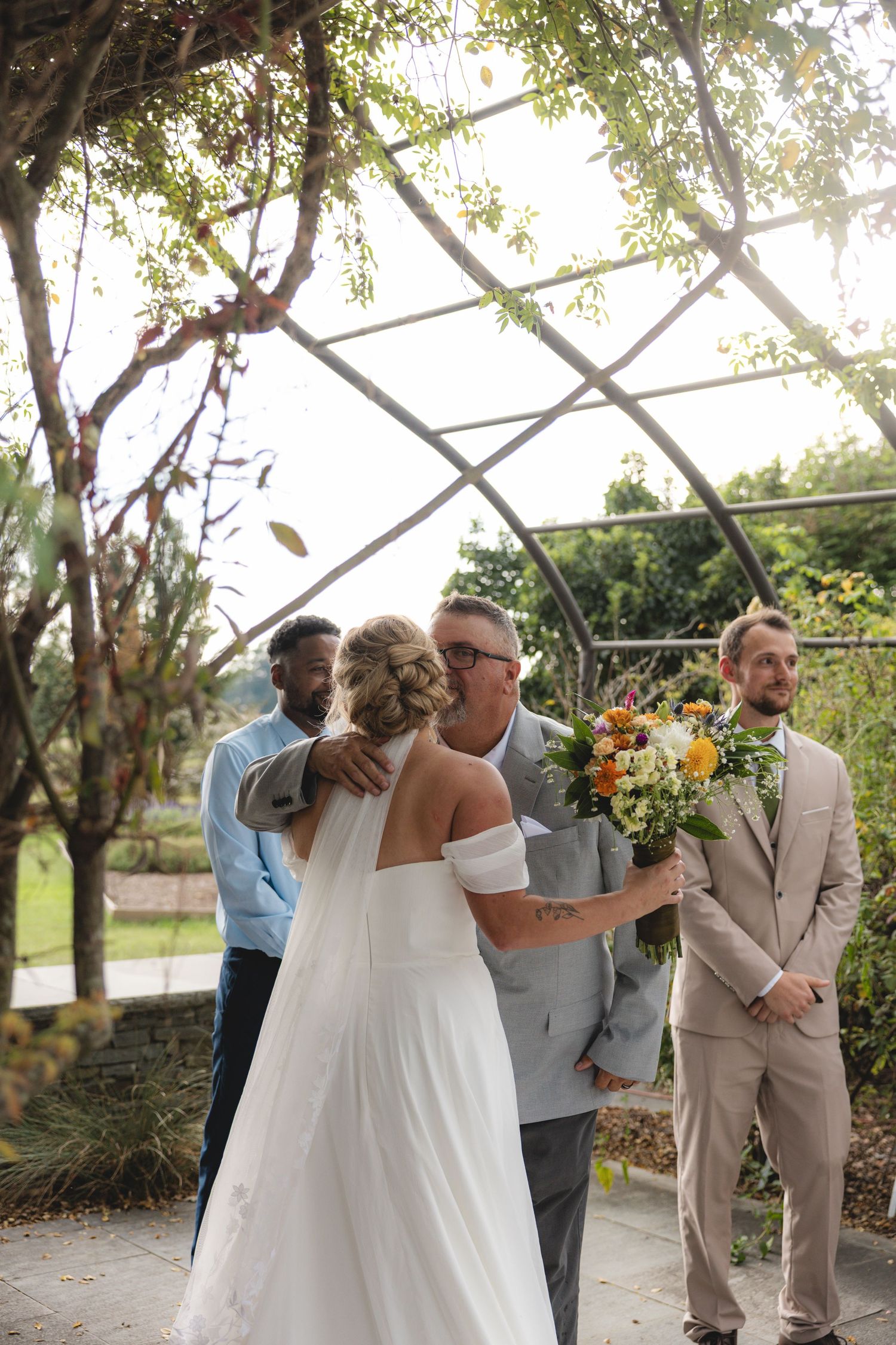 Bride in white gown receiving hug from father at outdoor wedding ceremony under garden arbor with groom waiting nearby.