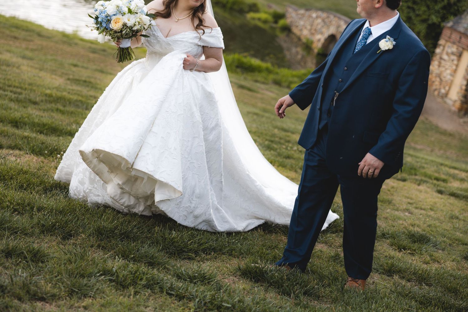 Bride in white ball gown and groom in navy suit walking on green grass at outdoor wedding ceremony.