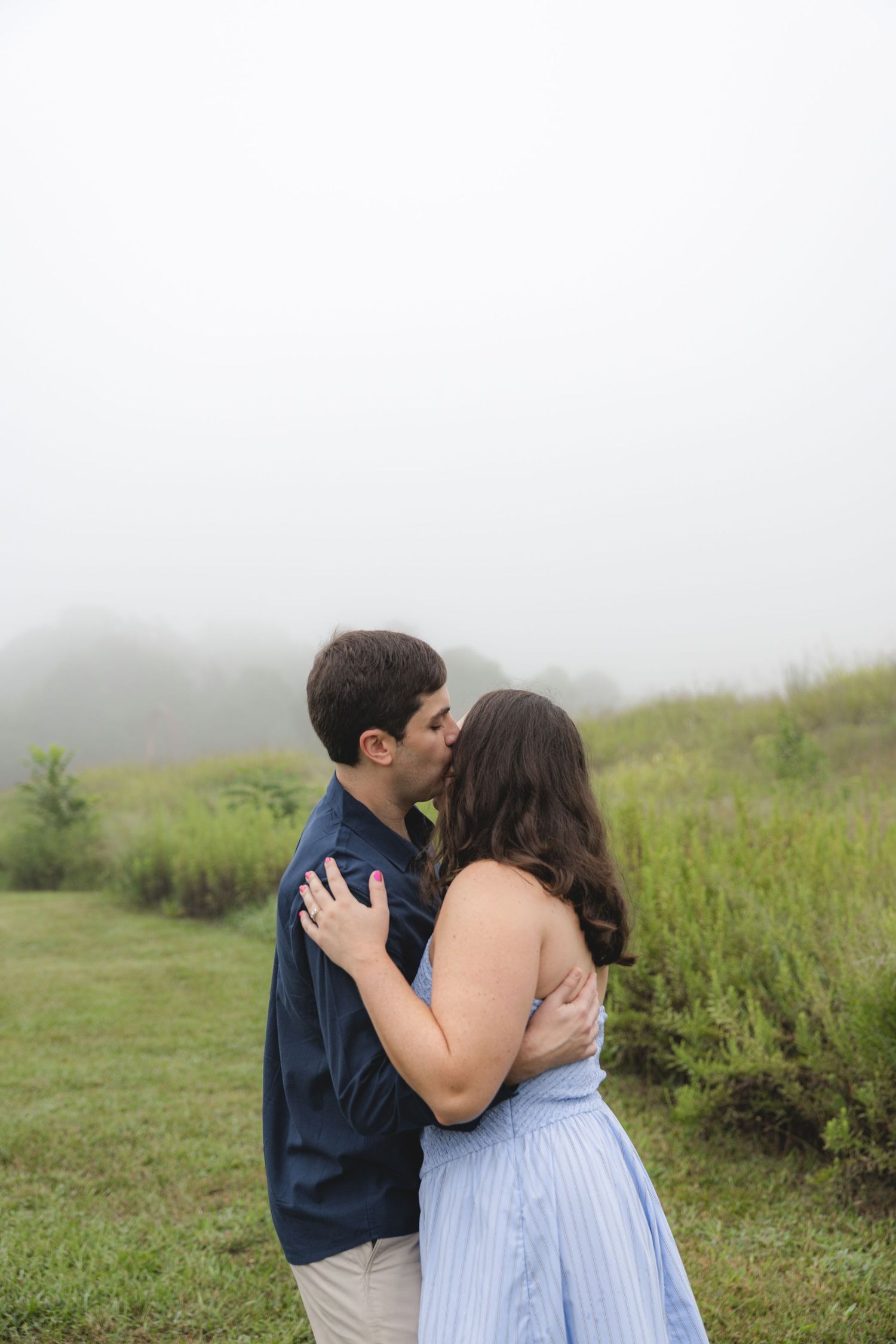 Couple sharing a romantic kiss in a misty green field, man in navy shirt embracing woman in light blue dress.