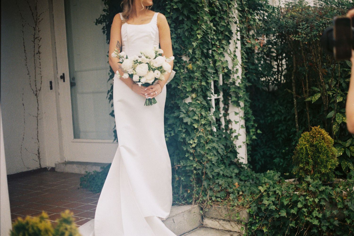 Bride in elegant white mermaid wedding gown holding white bouquet, standing in garden with ivy backdrop.