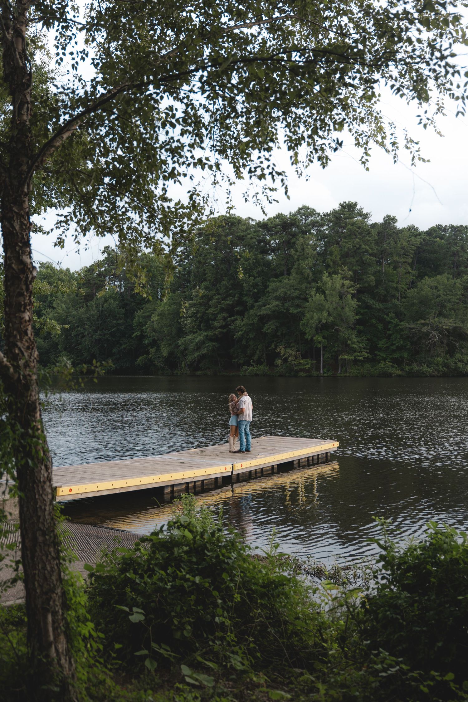 Couple embracing on a wooden dock surrounded by a serene lake and lush green trees.