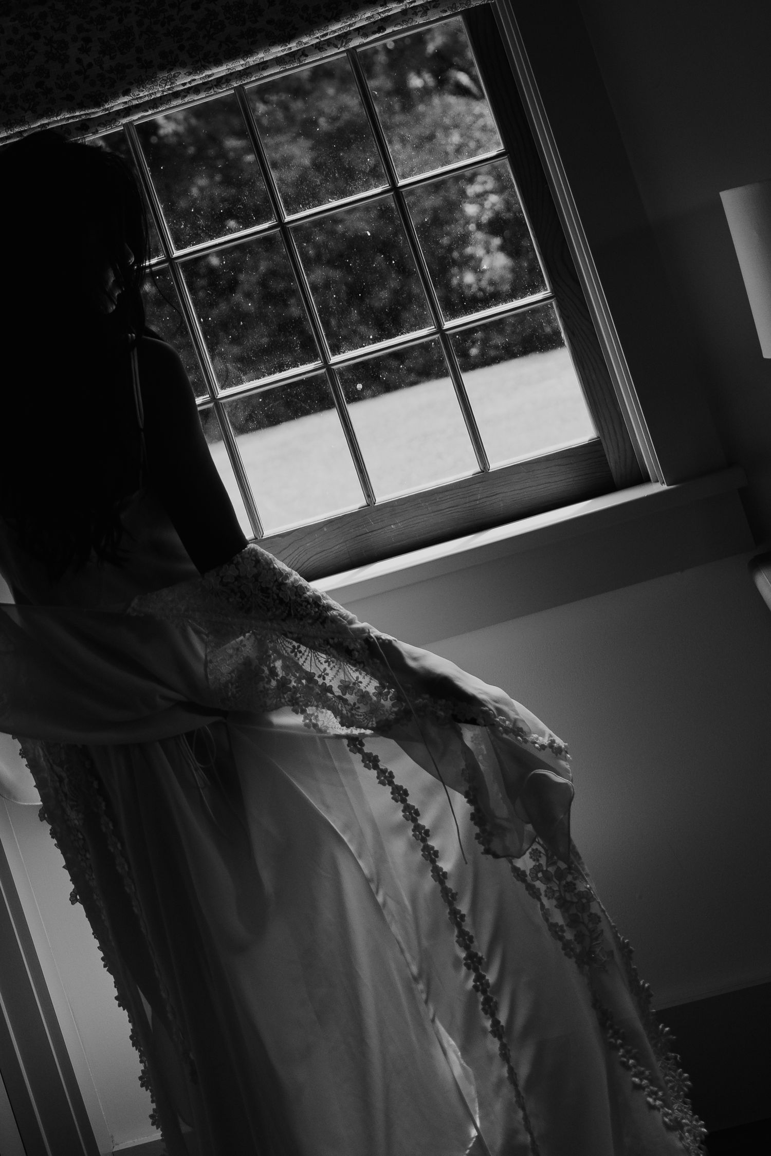 Black and white photo of a bride in lace wedding dress standing by a window with natural light.