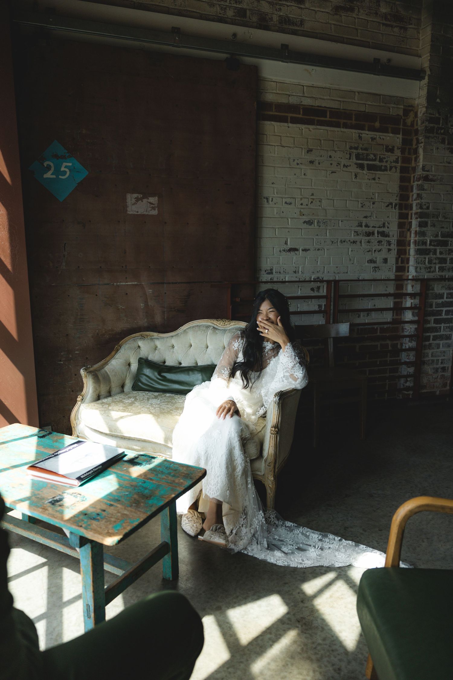A bride in a white lace gown sits on a vintage sofa in a rustic sunlit room with brick walls.