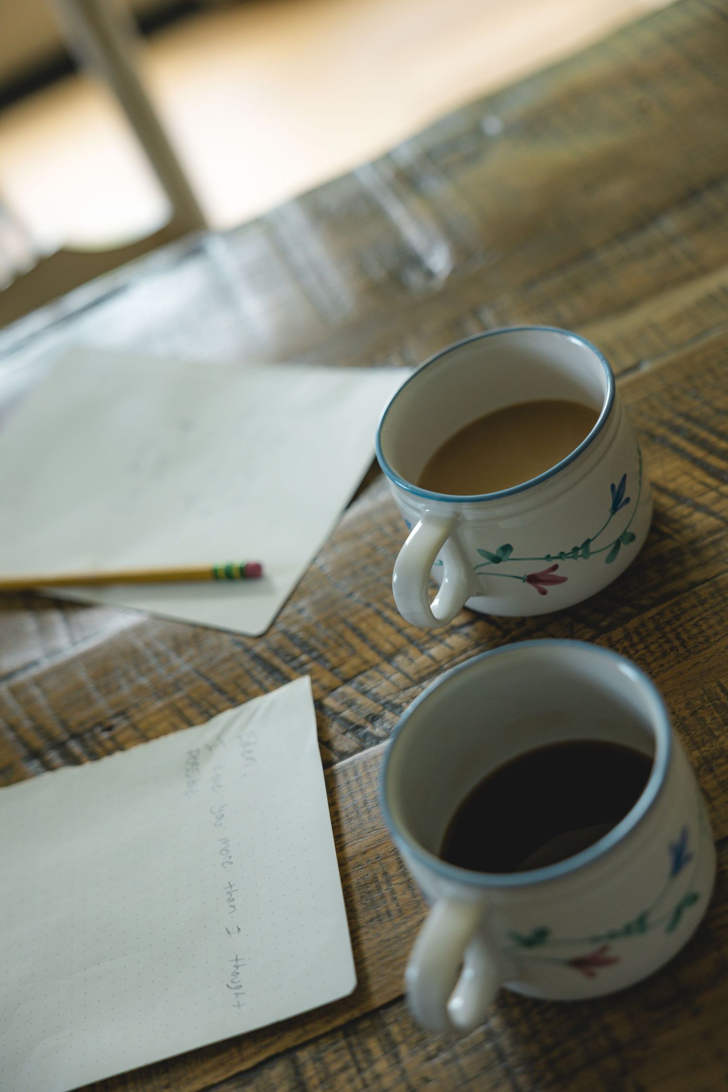 Two ceramic mugs of coffee on a wooden table with handwritten notes and a pencil nearby.