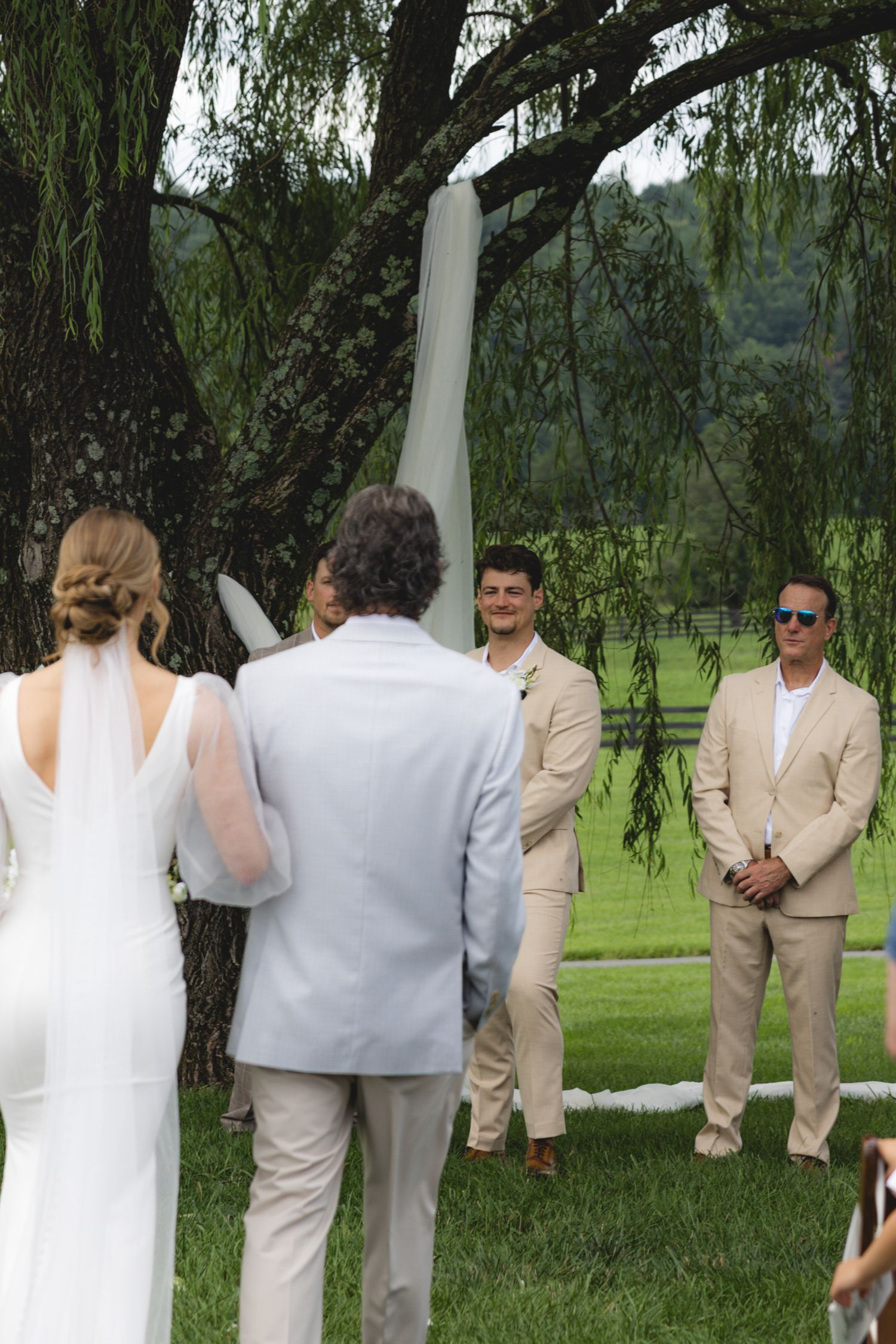 Outdoor wedding ceremony under a willow tree with bride in white gown facing groom in tan suit with groomsmen.