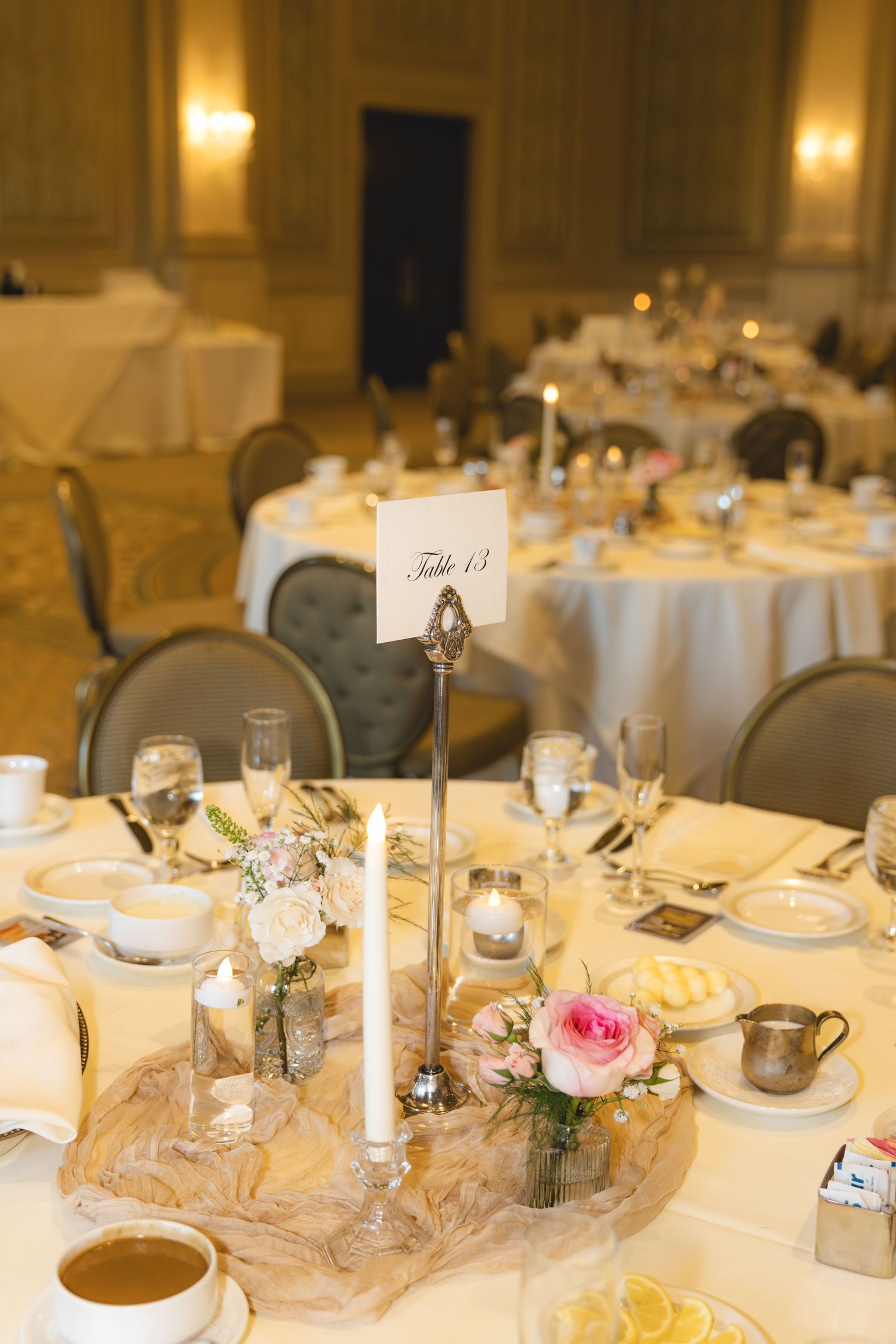 Wedding reception table 13 setting with pink roses, white candles, burlap centerpiece, and fine china in elegant ballroom.