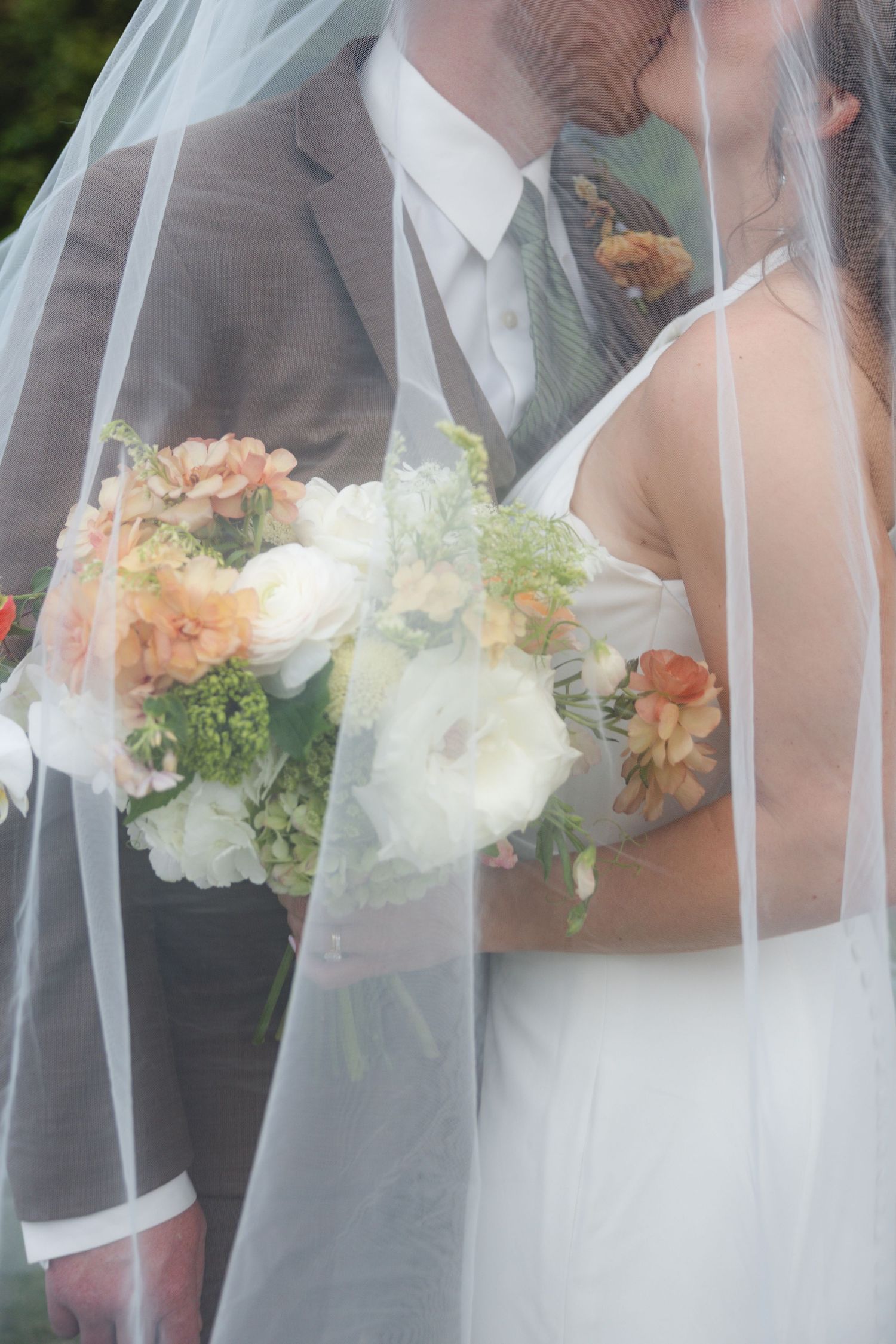 Bride and groom kissing under a sheer veil while holding a peach and white floral wedding bouquet.