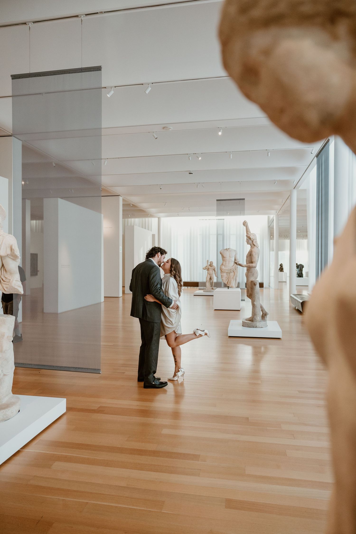 A couple embraces in an art museum gallery surrounded by classical sculptures.