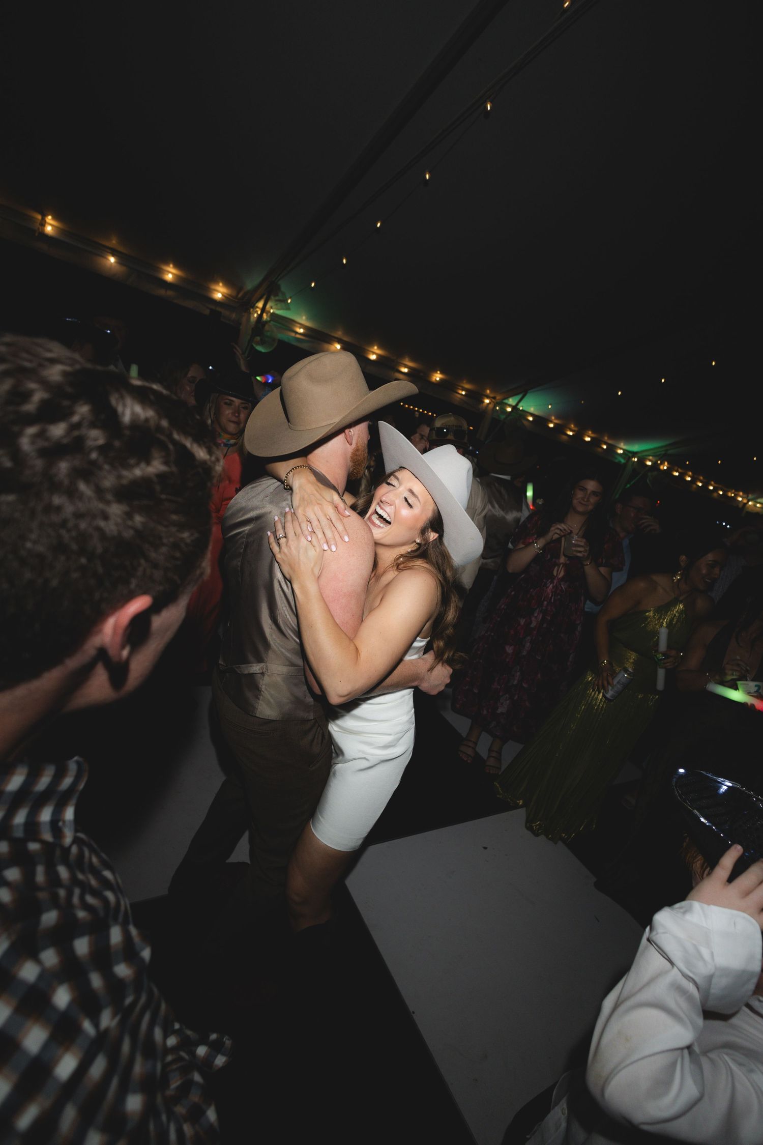 Couple dancing joyfully under string lights at a lively wedding reception, both wearing cowboy hats.