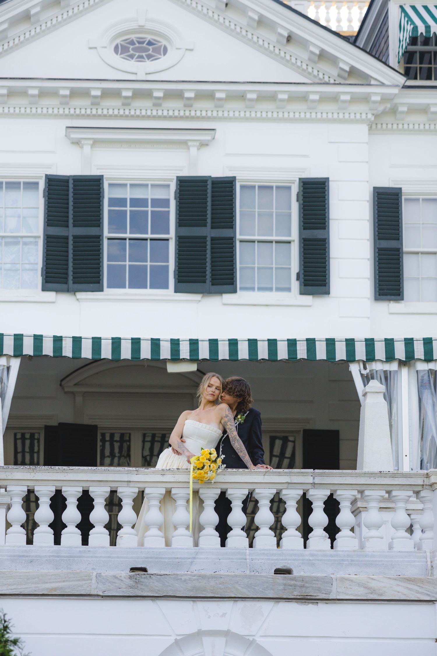 Bride and groom share a romantic moment on a white balcony of an elegant estate with green shutters.