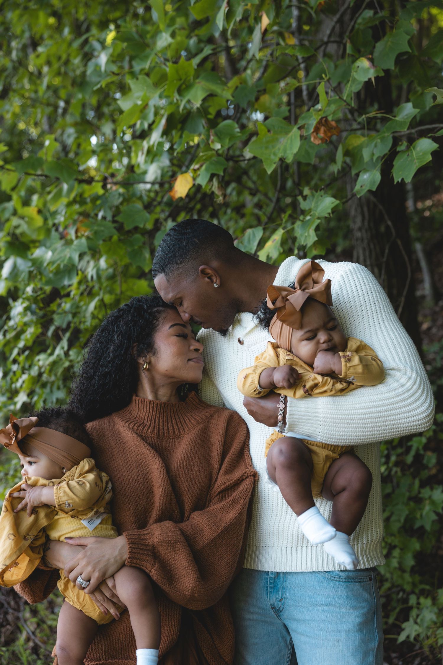 A loving Black couple shares a tender moment outdoors in autumn, each cradling a baby dressed in yellow amid lush green foliage.