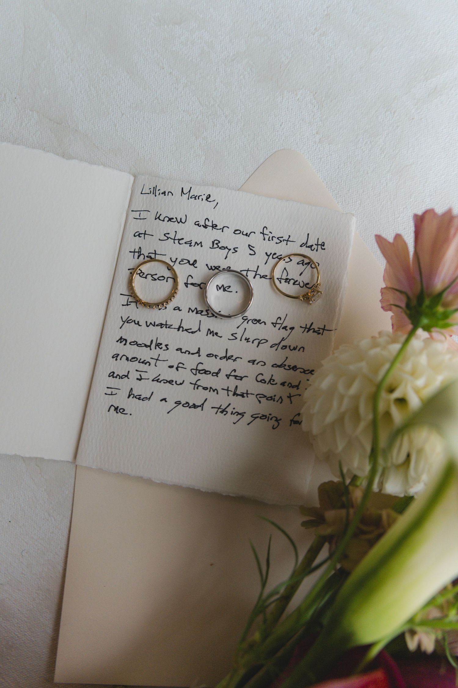Handwritten wedding vow letter to Lilian Marie with three gold rings resting on the page, beside pink and white flowers.