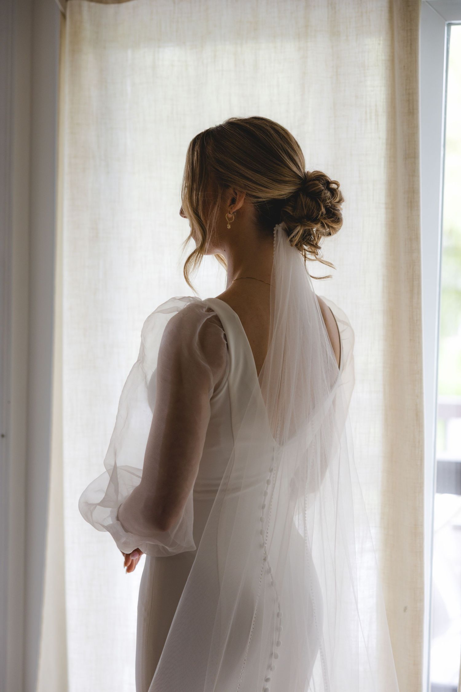 Bride in white gown with sheer sleeves and veil, elegant updo, standing by window in soft natural light.