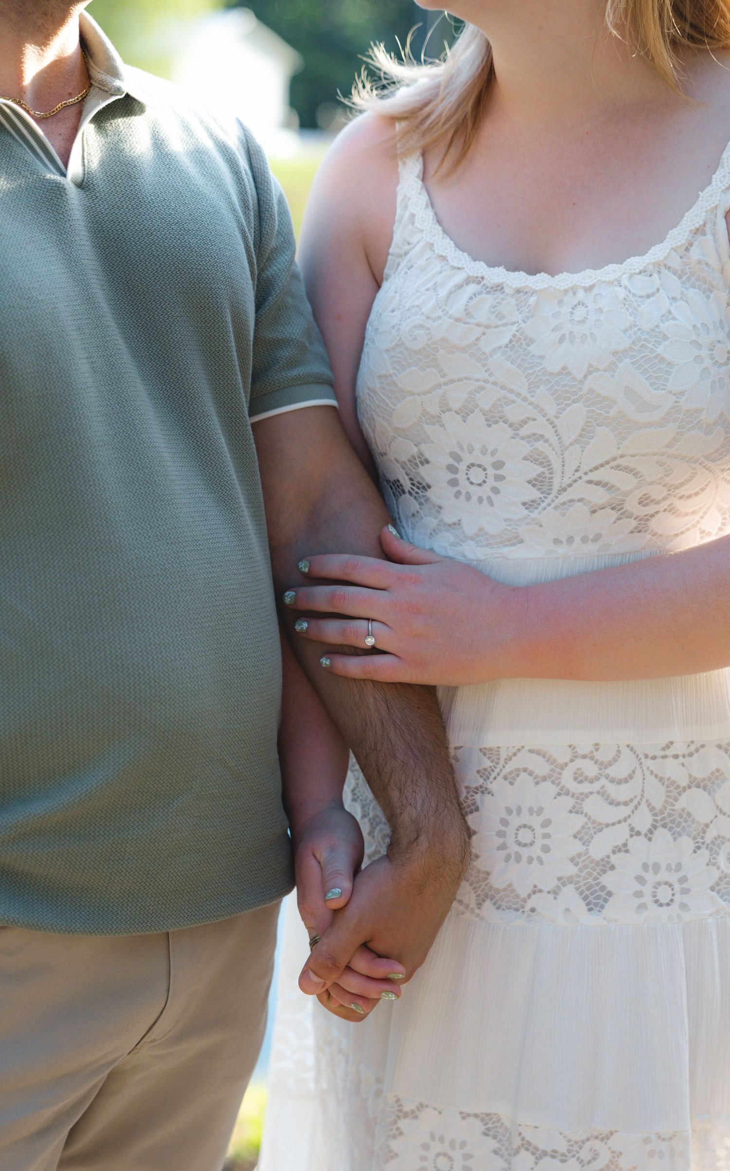 Couple holding hands, woman in white lace dress with engagement ring, man in gray polo shirt, outdoor setting.