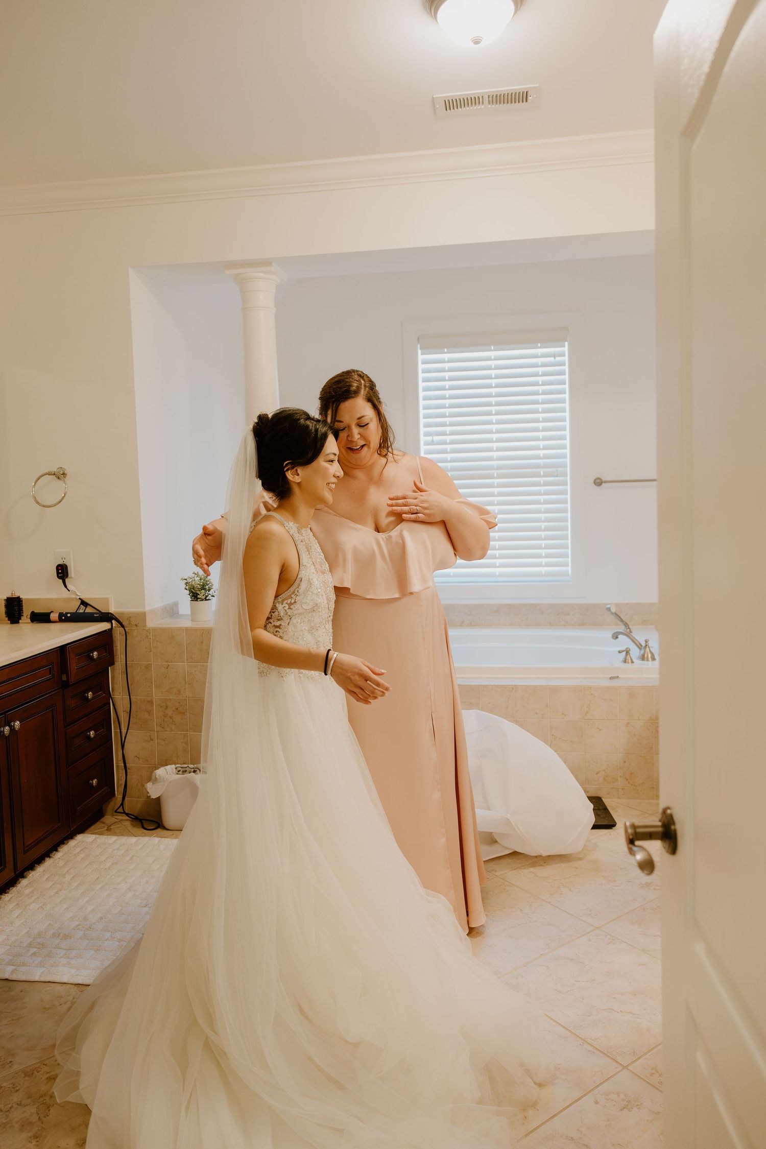 Bride in lace A-line wedding gown shares a tender moment with bridesmaid in blush dress in bathroom.
