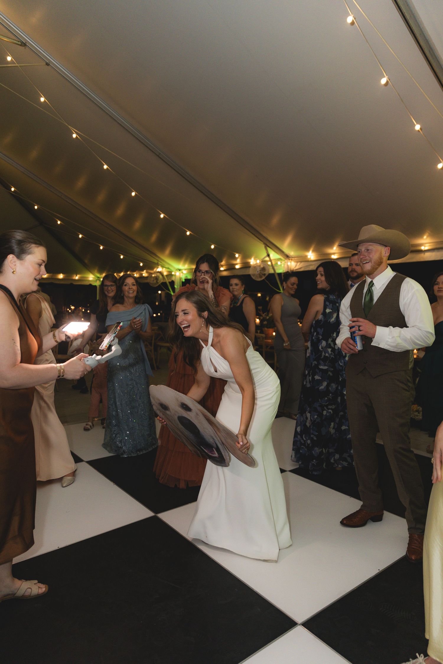 Laughing bride in white dress dances with guests on checkered floor at lively wedding reception.