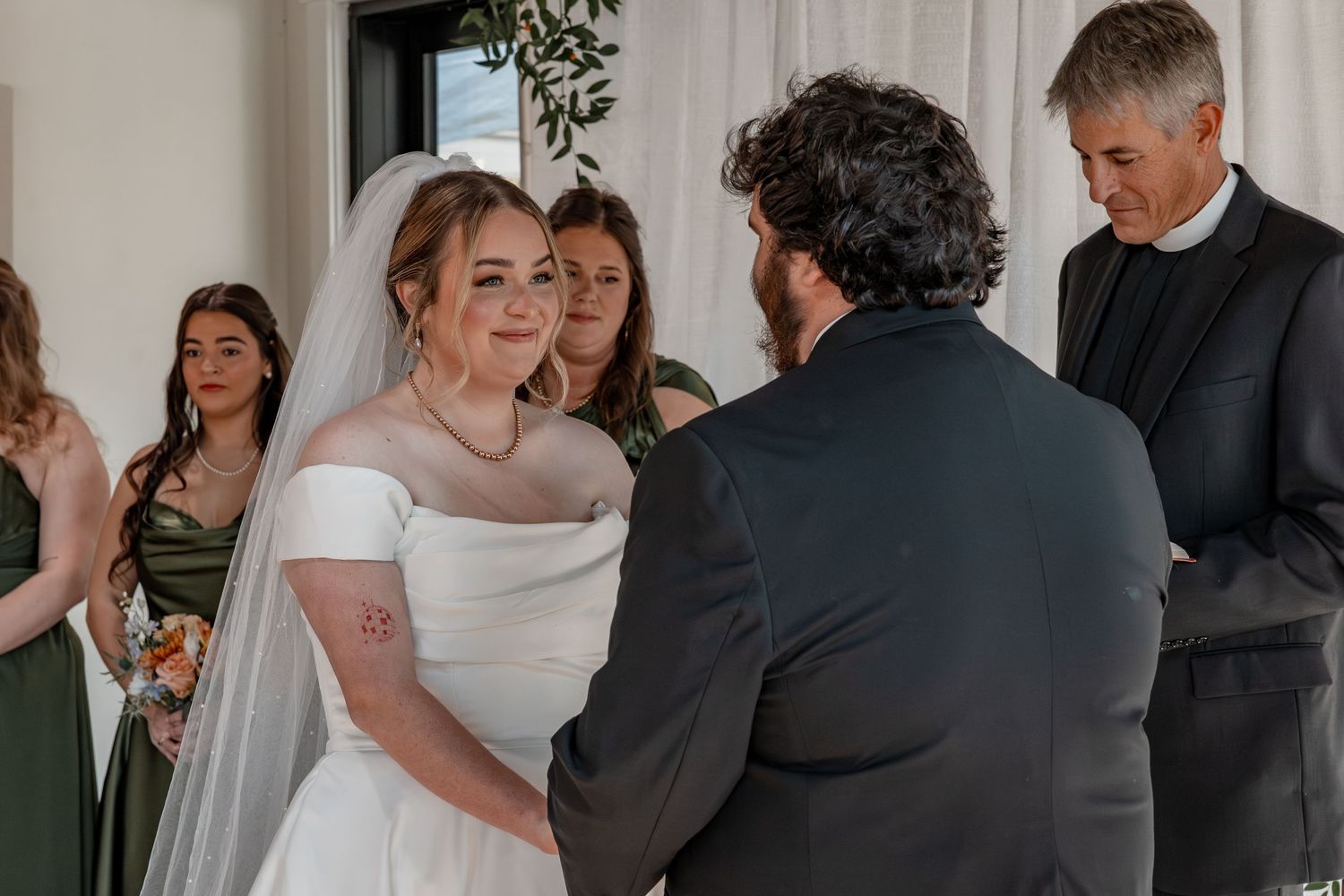 Bride in white gown exchanging vows with groom at indoor wedding ceremony with bridesmaids in sage green.