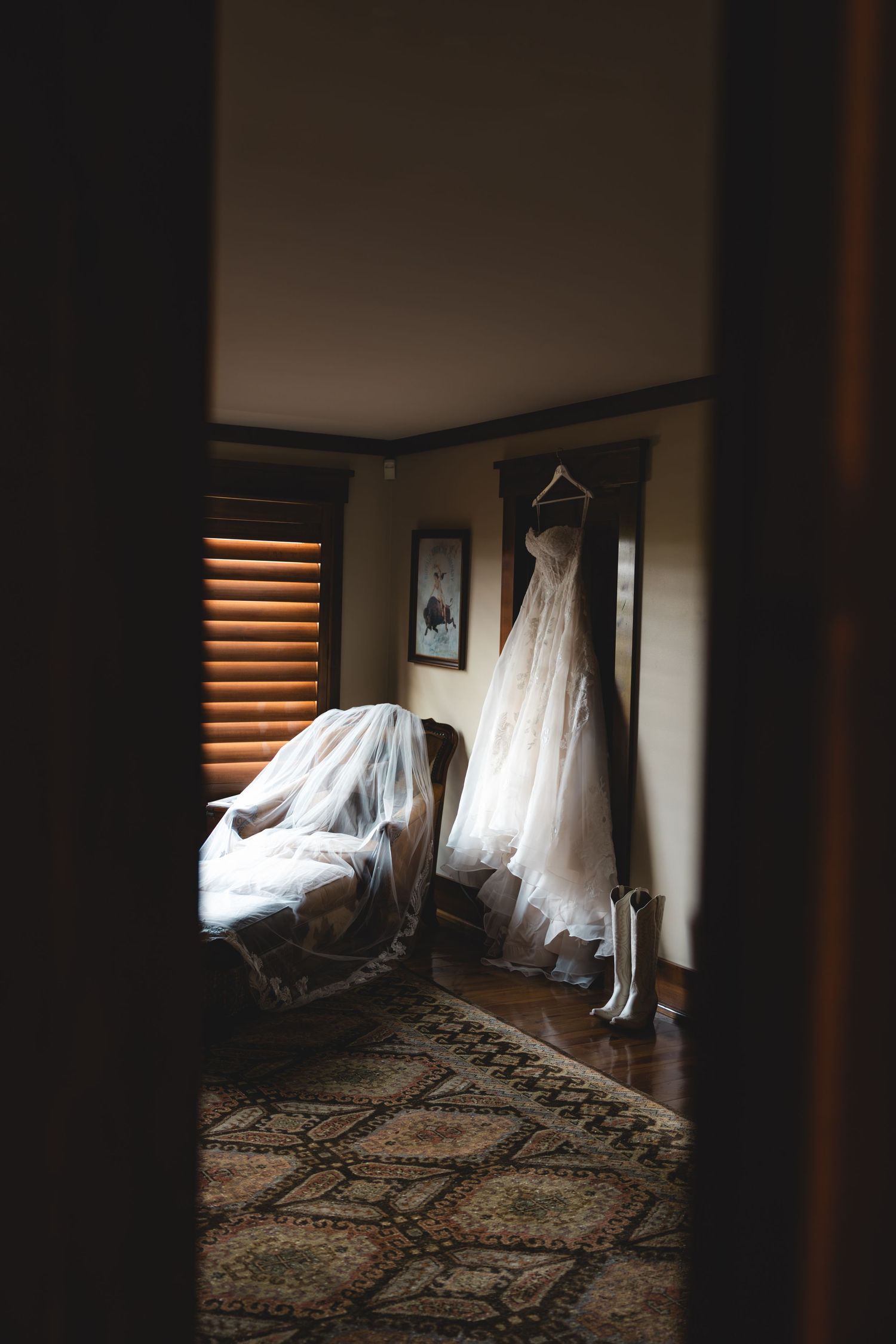Wedding dress hanging in a dimly lit room with bridal veil draped over a chair and white boots below.