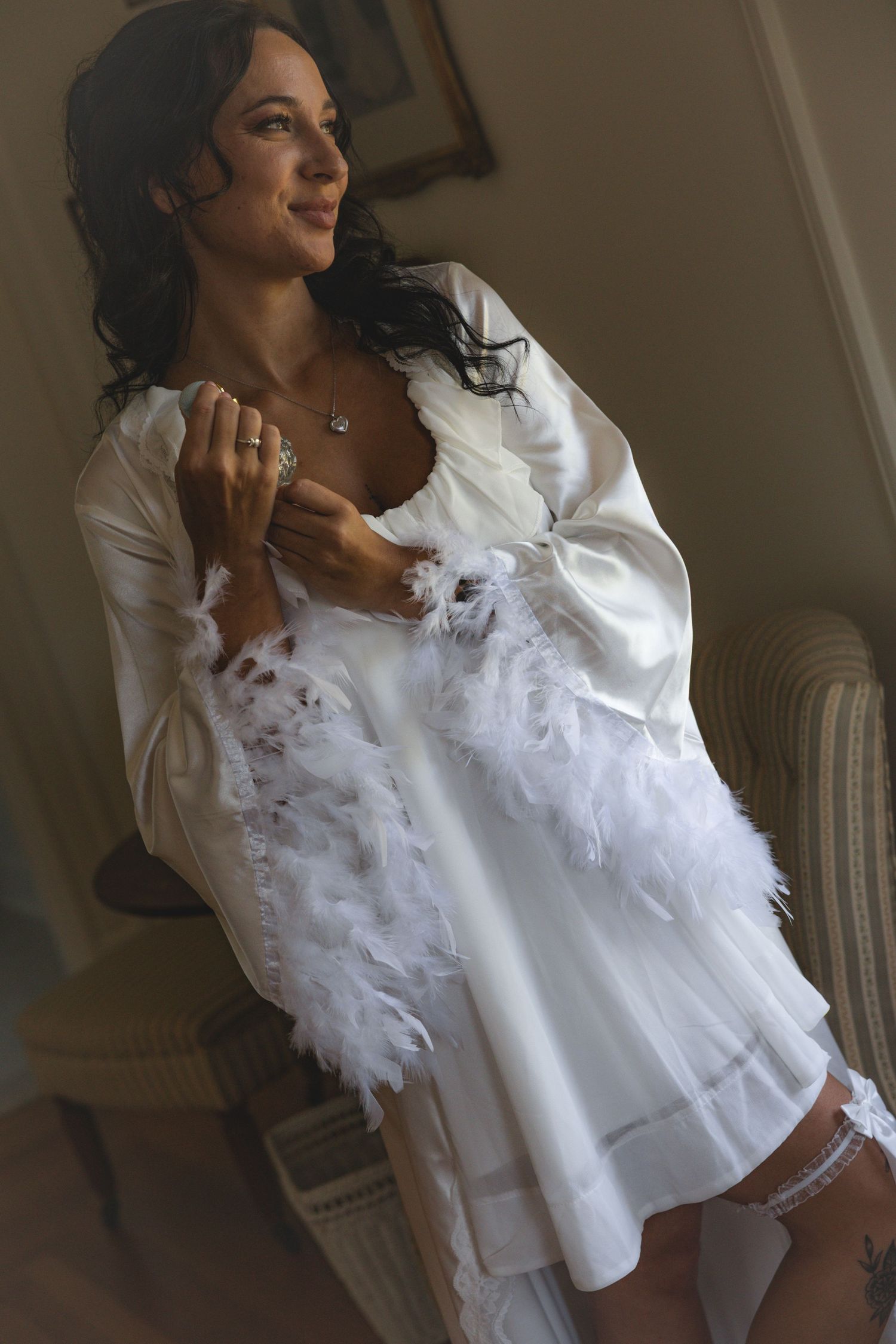 A woman in a white satin robe and feather boa poses elegantly in a warmly lit room.