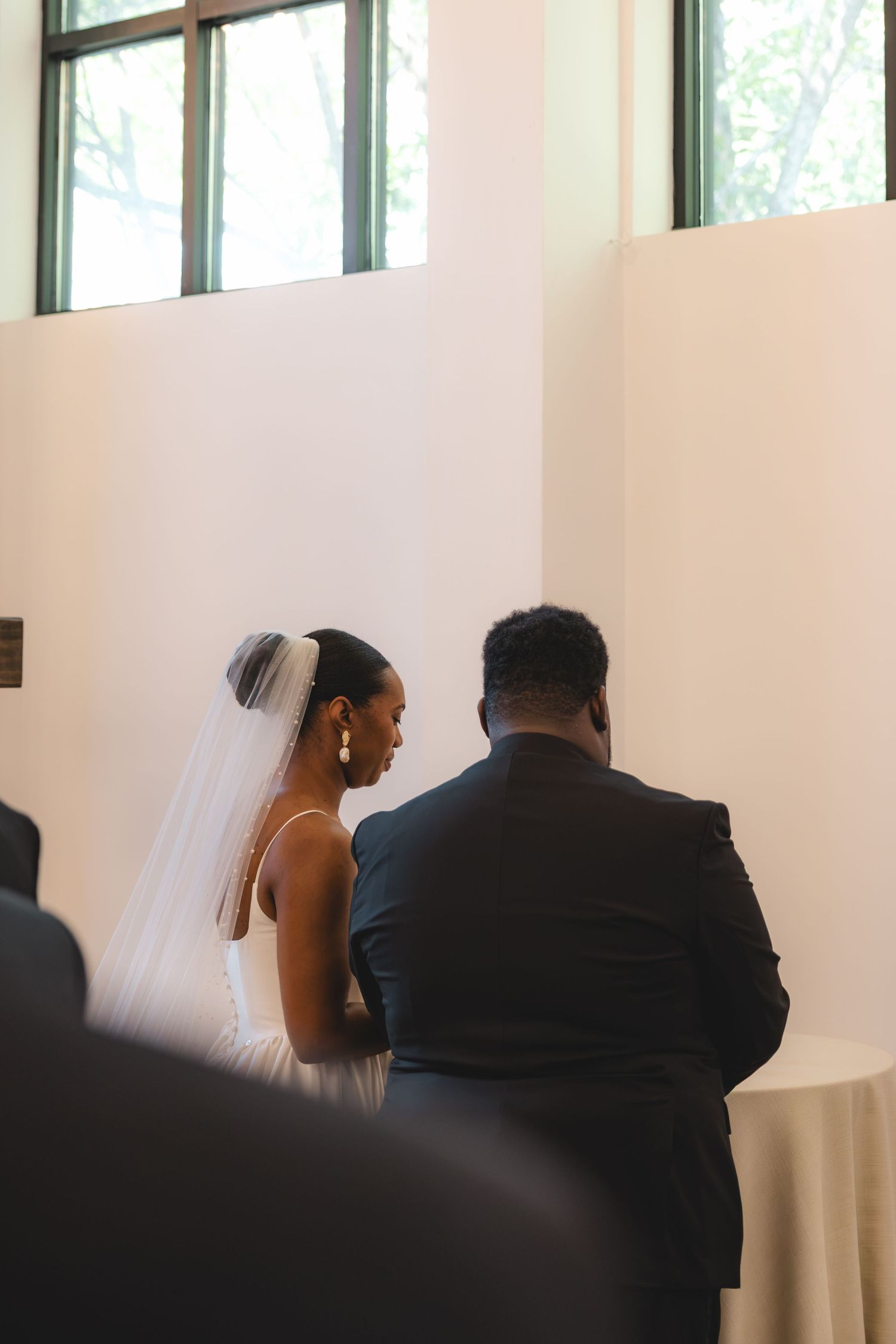 A bride in a white gown and veil stands with a groom in a navy suit during an intimate wedding ceremony indoors.