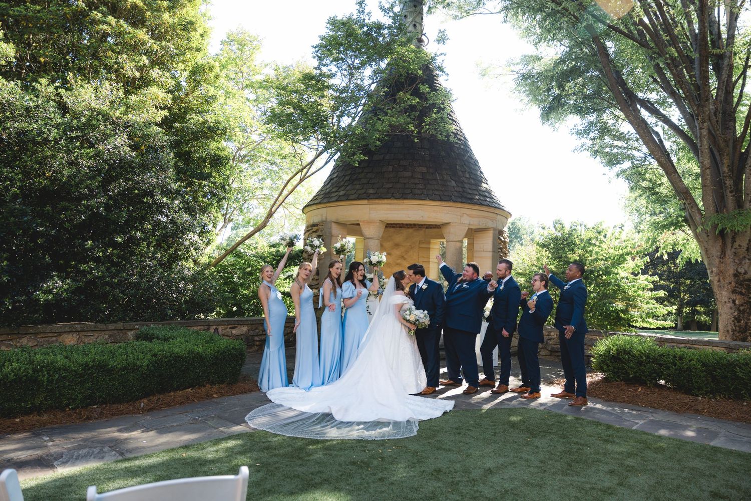 Wedding party poses near a gazebo in a lush garden, bride in white gown with bridesmaids in blue dresses.
