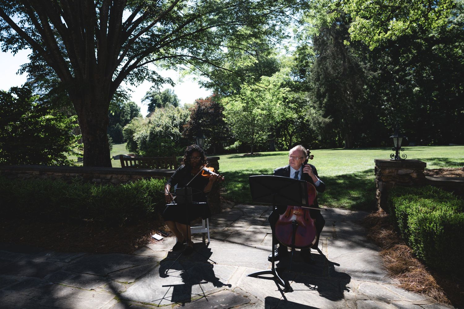 Two musicians playing cellos outdoors in a lush green garden park on a sunny day.