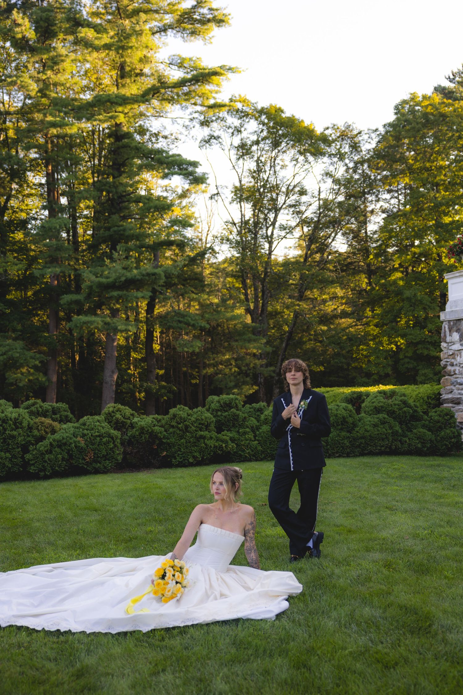 Bride in white gown sits on green lawn holding yellow bouquet while groom in black suit stands behind her outdoors.