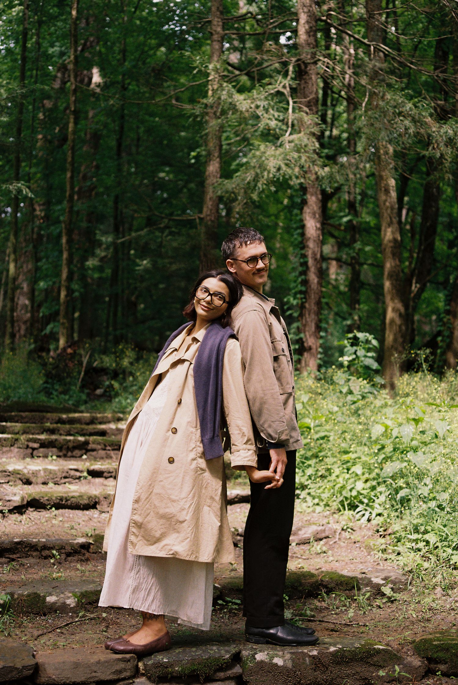 Couple in beige trench coats posing back-to-back on stone steps in a lush green forest setting.