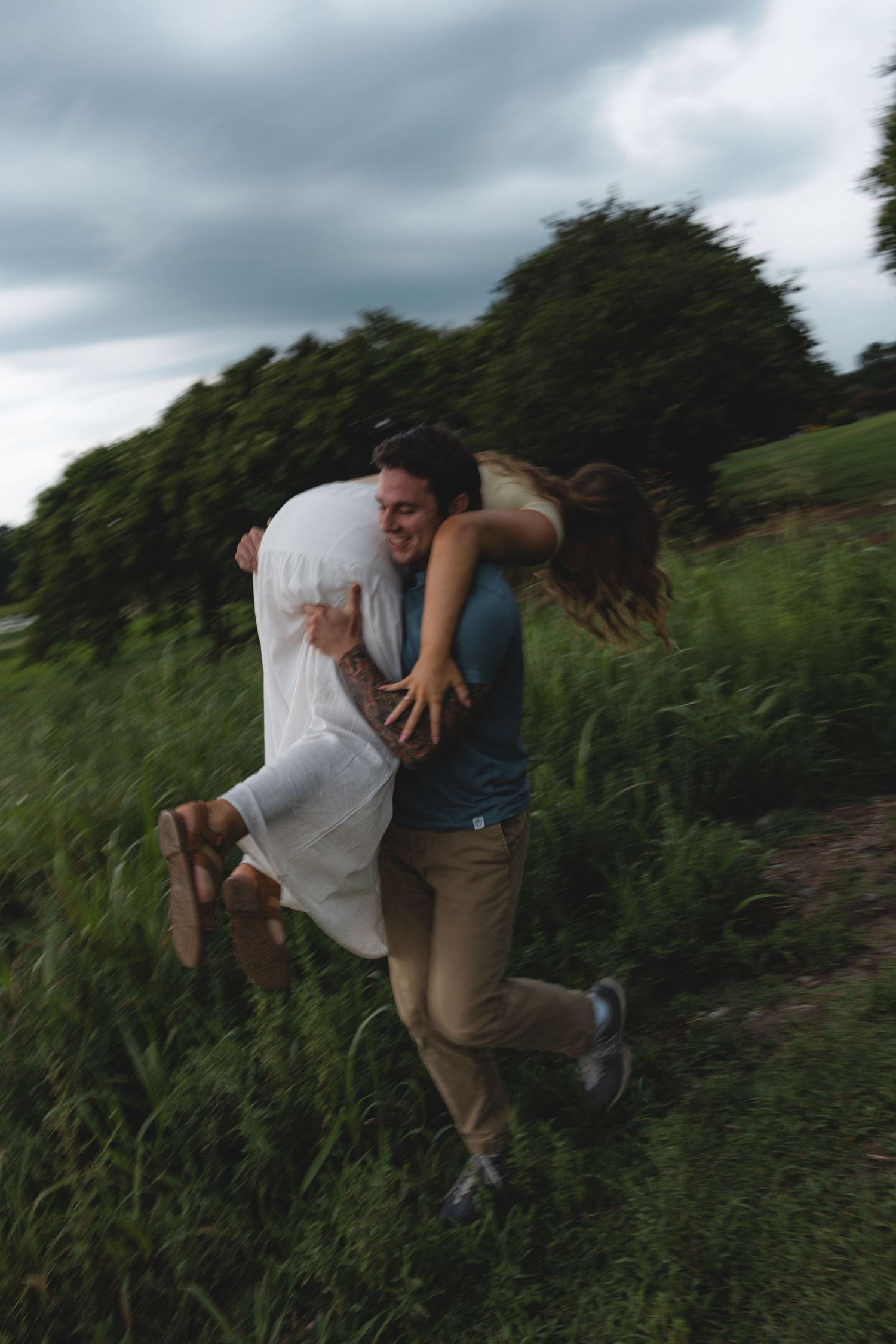 A joyful couple embraces outdoors, man lifting woman in white dress, surrounded by lush green fields under cloudy skies.