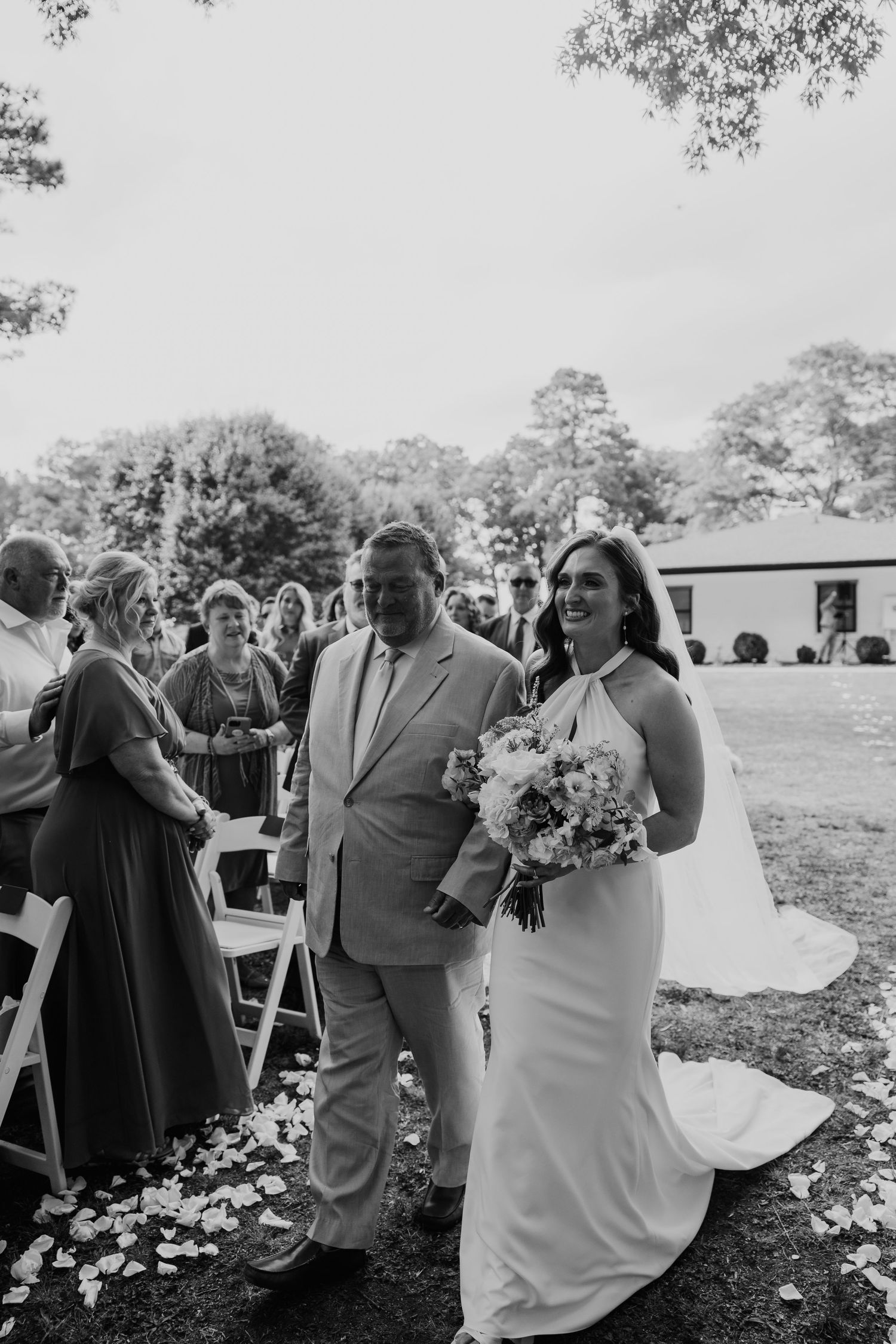 Black and white photo of a smiling bride in a halter gown walking down the aisle with her father at an outdoor wedding.