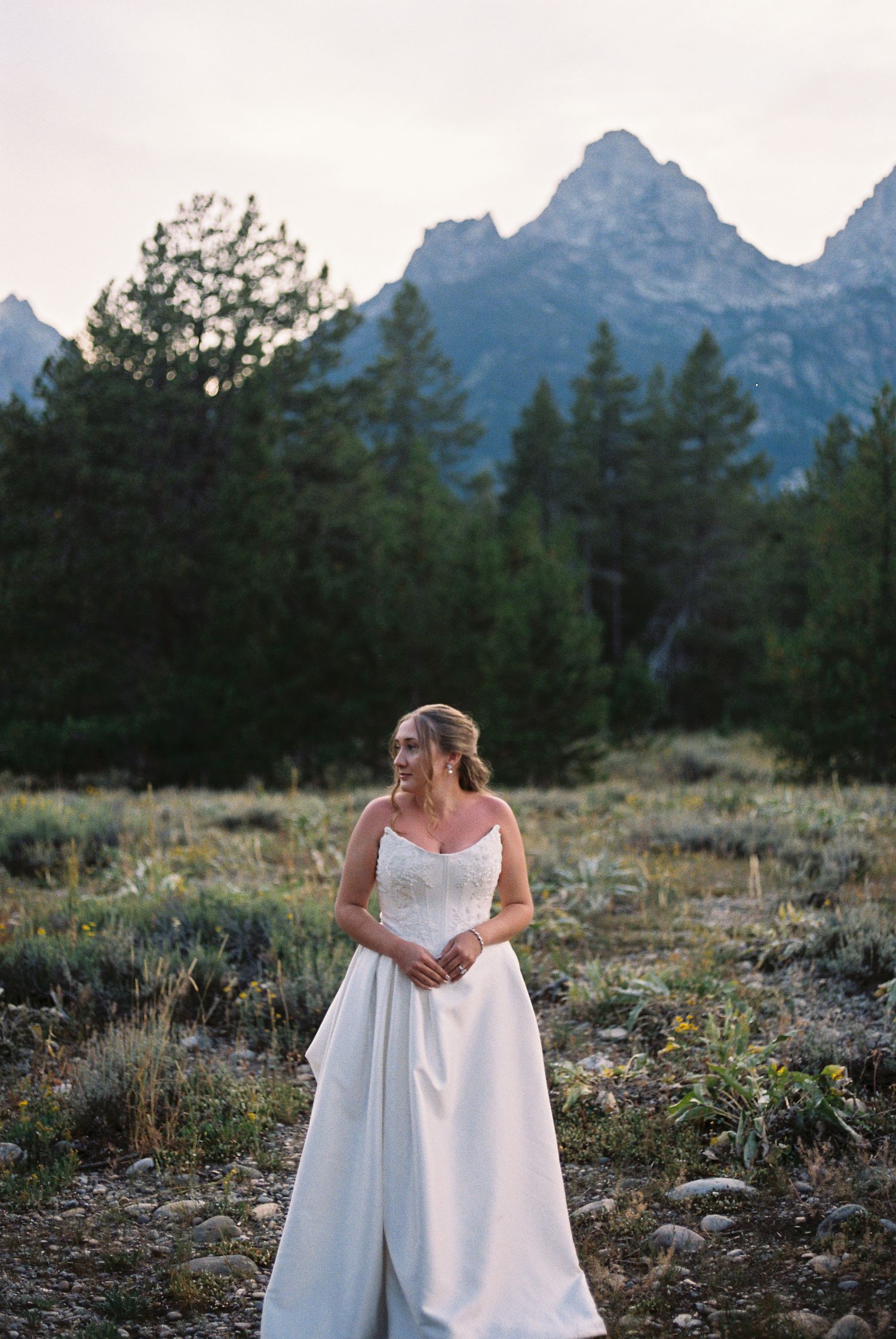 Bride in strapless lace ballgown stands in mountain meadow with Grand Teton peaks and pine trees in background.