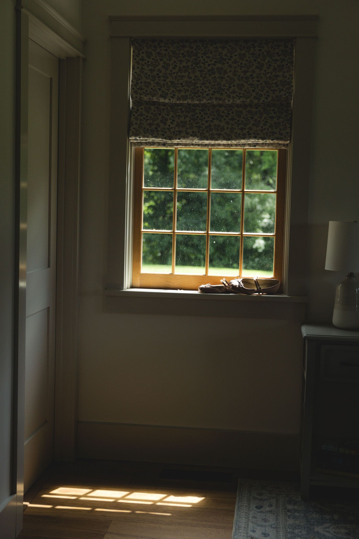 Sunlight streams through a small nine-pane window with floral roman shade, casting grid shadows on hardwood floor in cozy room.