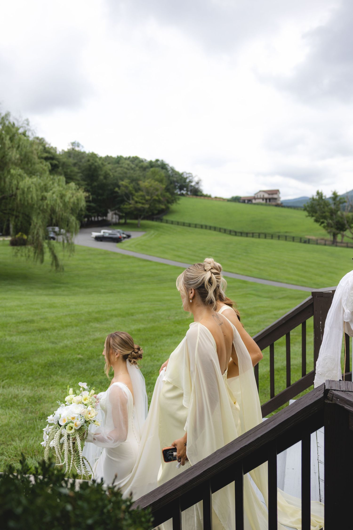Bride and bridesmaid descend wooden stairs at outdoor wedding venue with lush green hills and willow trees in background.