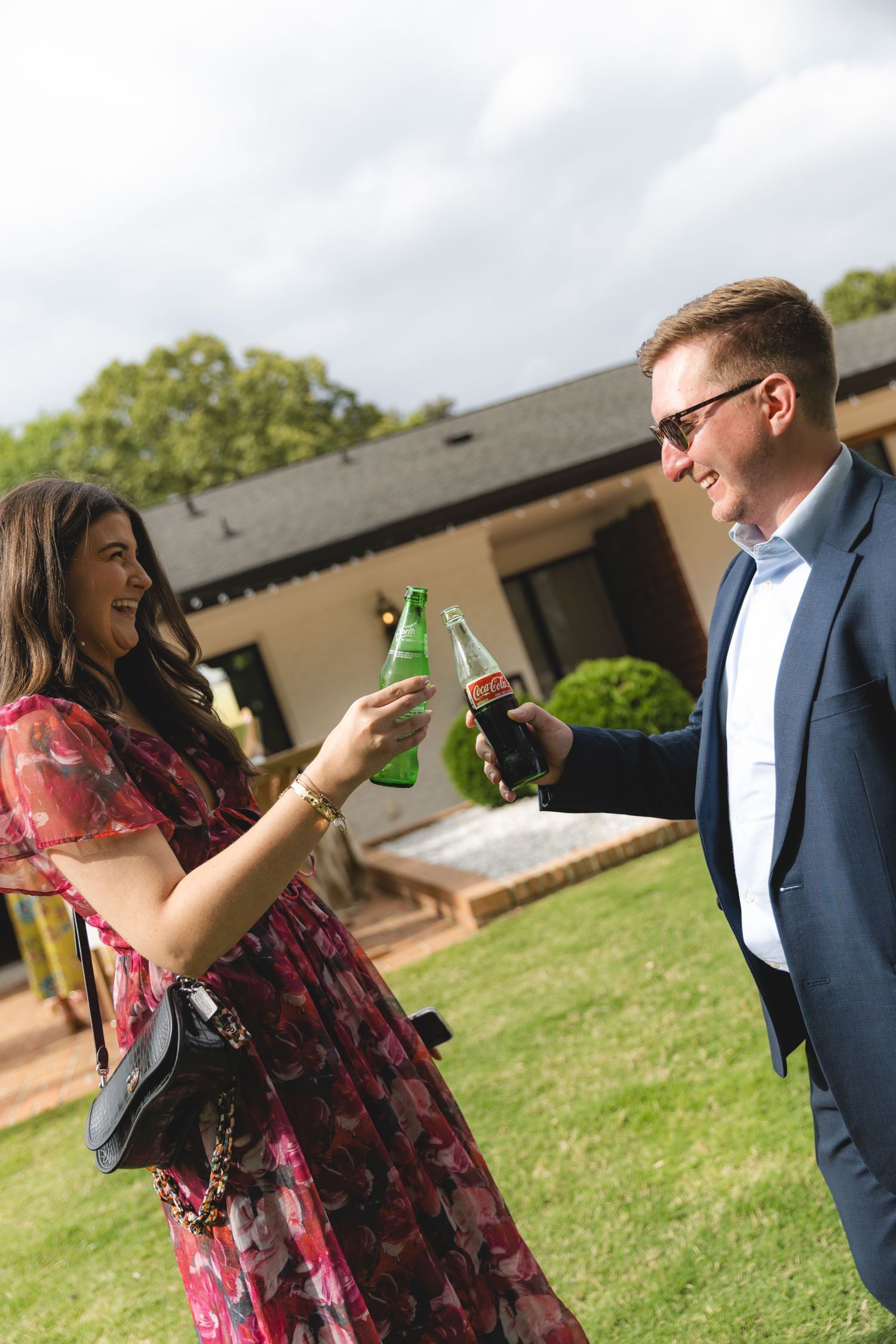 A couple toasting with glass bottles outdoors at a garden party, woman in floral dress, man in navy suit.