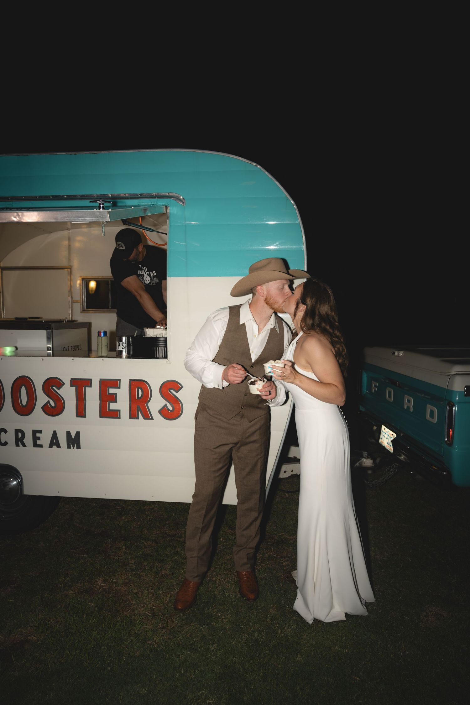 Newlywed couple kissing beside a teal Roosters Ice Cream trailer at an outdoor wedding reception at night.