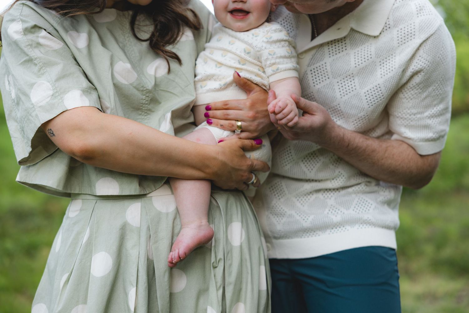 Parents in coordinating outfits hold their smiling baby together in a green outdoor setting.