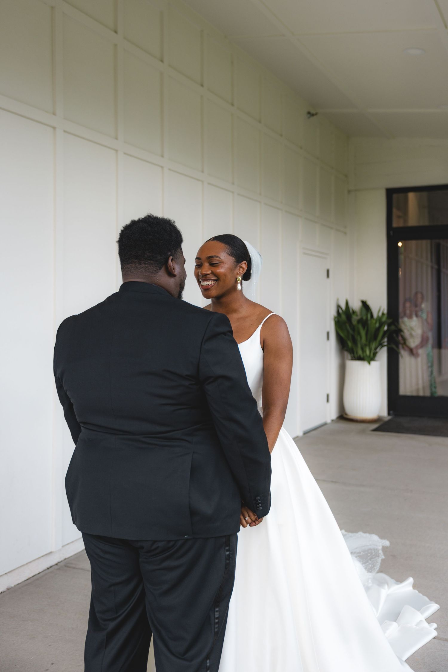 Smiling bride in a white gown holds hands with groom in black tuxedo during outdoor first look moment.