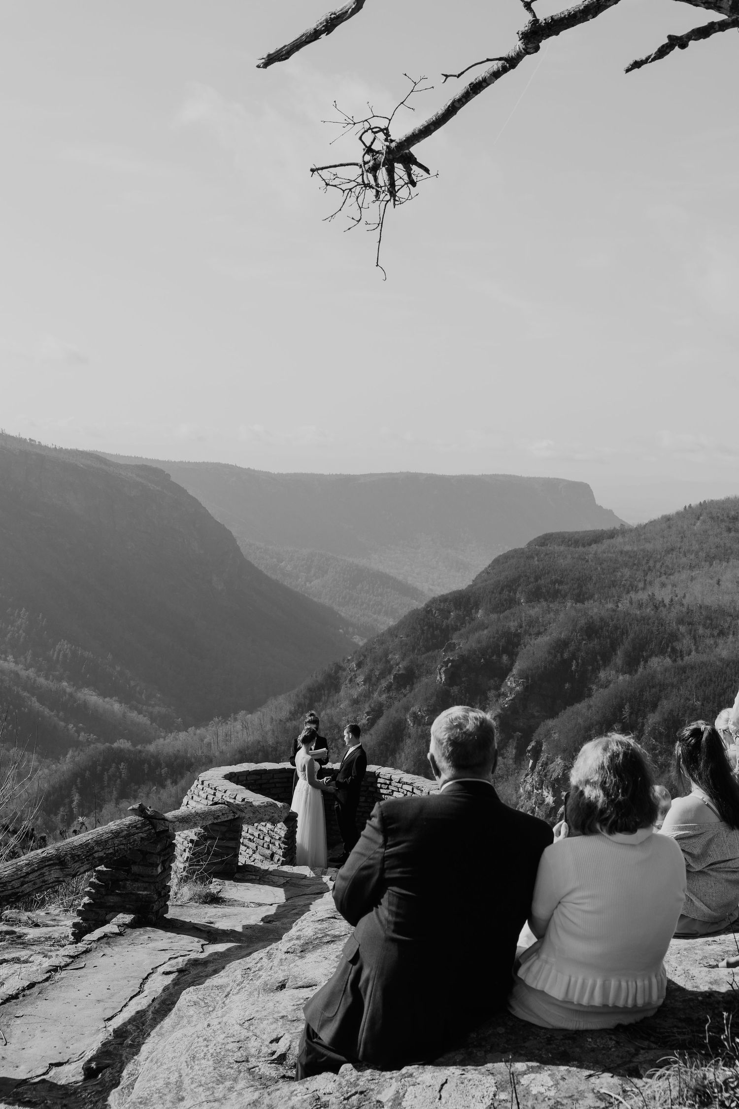 Wedding ceremony on mountain overlook with guests seated on stone wall and stunning valley views in black and white.