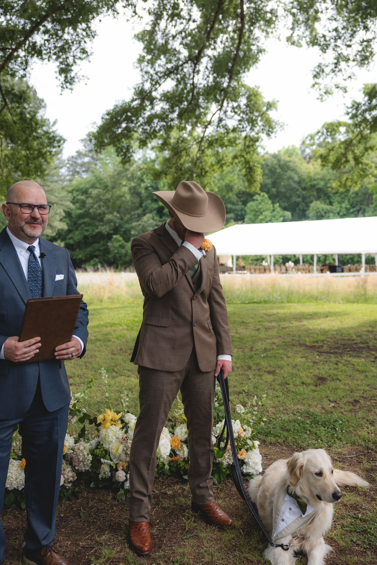 Groom in brown suit and cowboy hat crying at outdoor wedding ceremony with dog and officiant nearby.