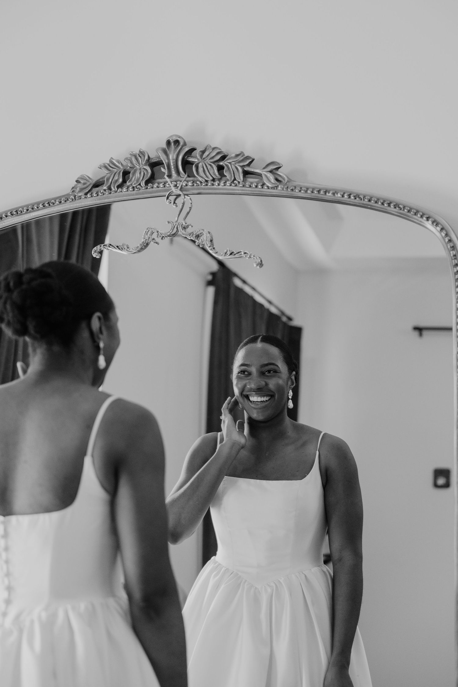 Black and white portrait of a smiling bride in white dress adjusting her earring in an ornate mirror.