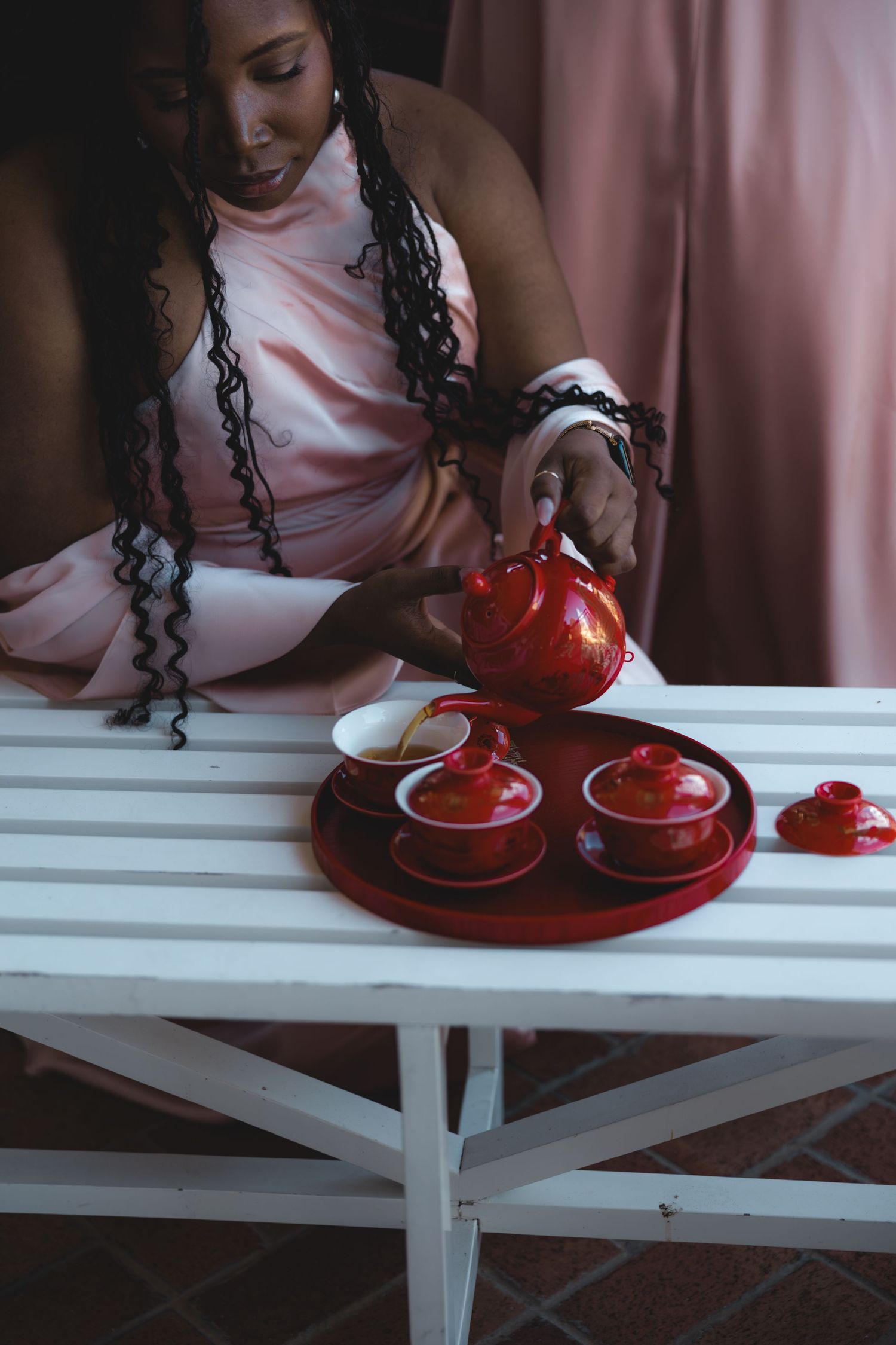 Bridesmaid in blush pink dress pouring tea from red Chinese teapot into cups on white bench.