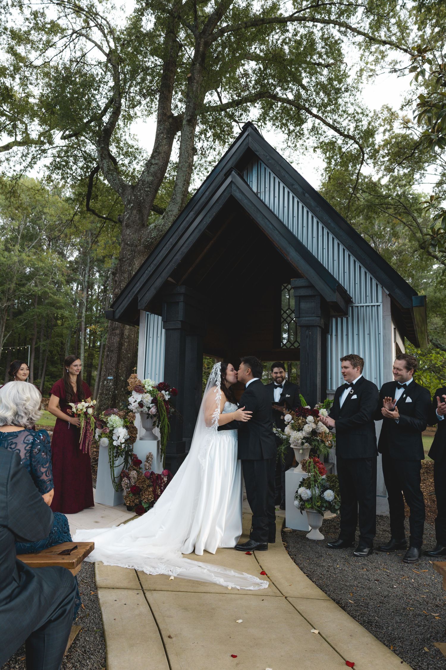 Bride in a long white gown and veil kisses the groom outside a rustic chapel as groomsmen and guests applaud in a wooded setting.