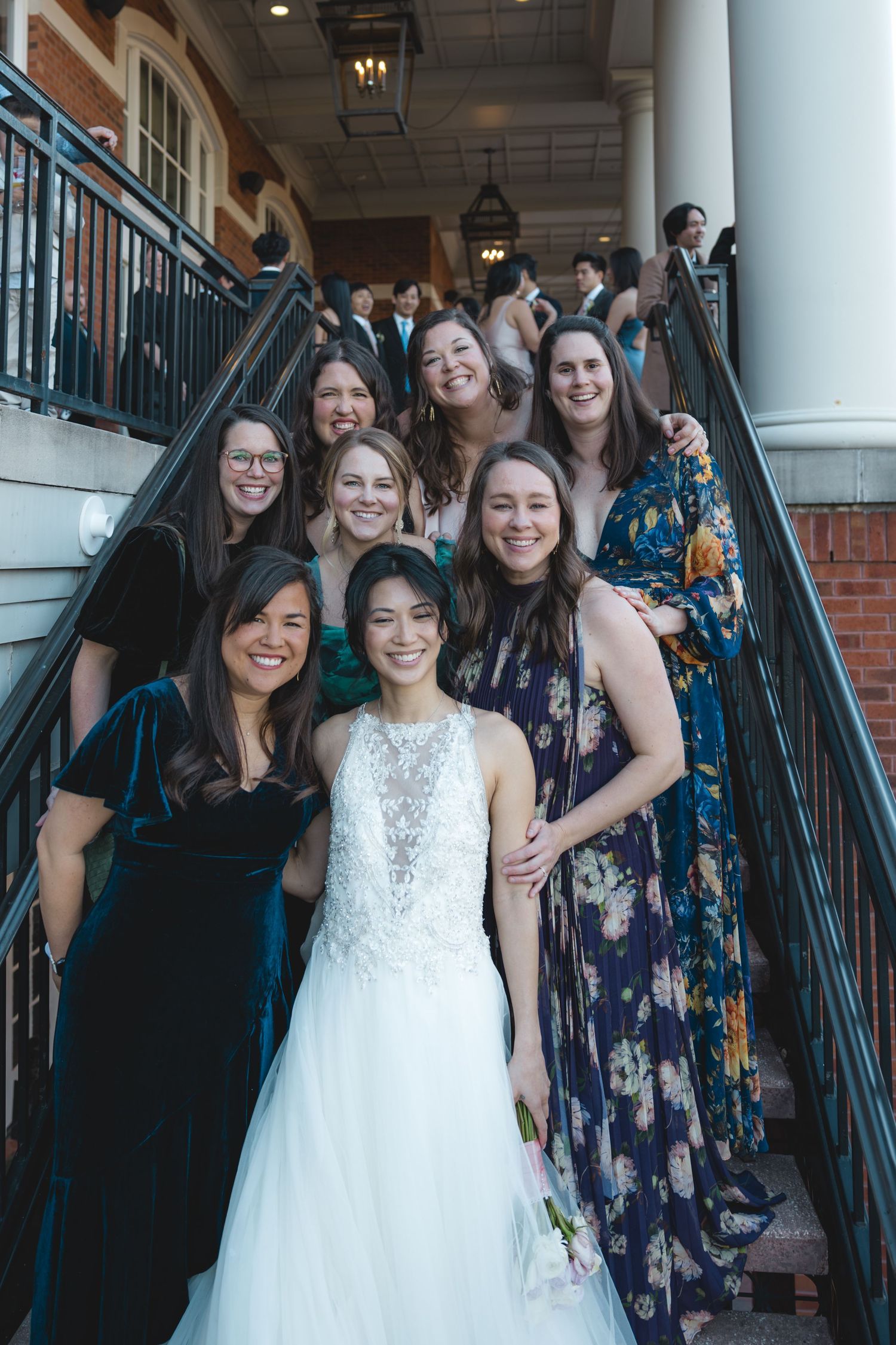 Bride surrounded by smiling female guests on staircase at elegant wedding reception.