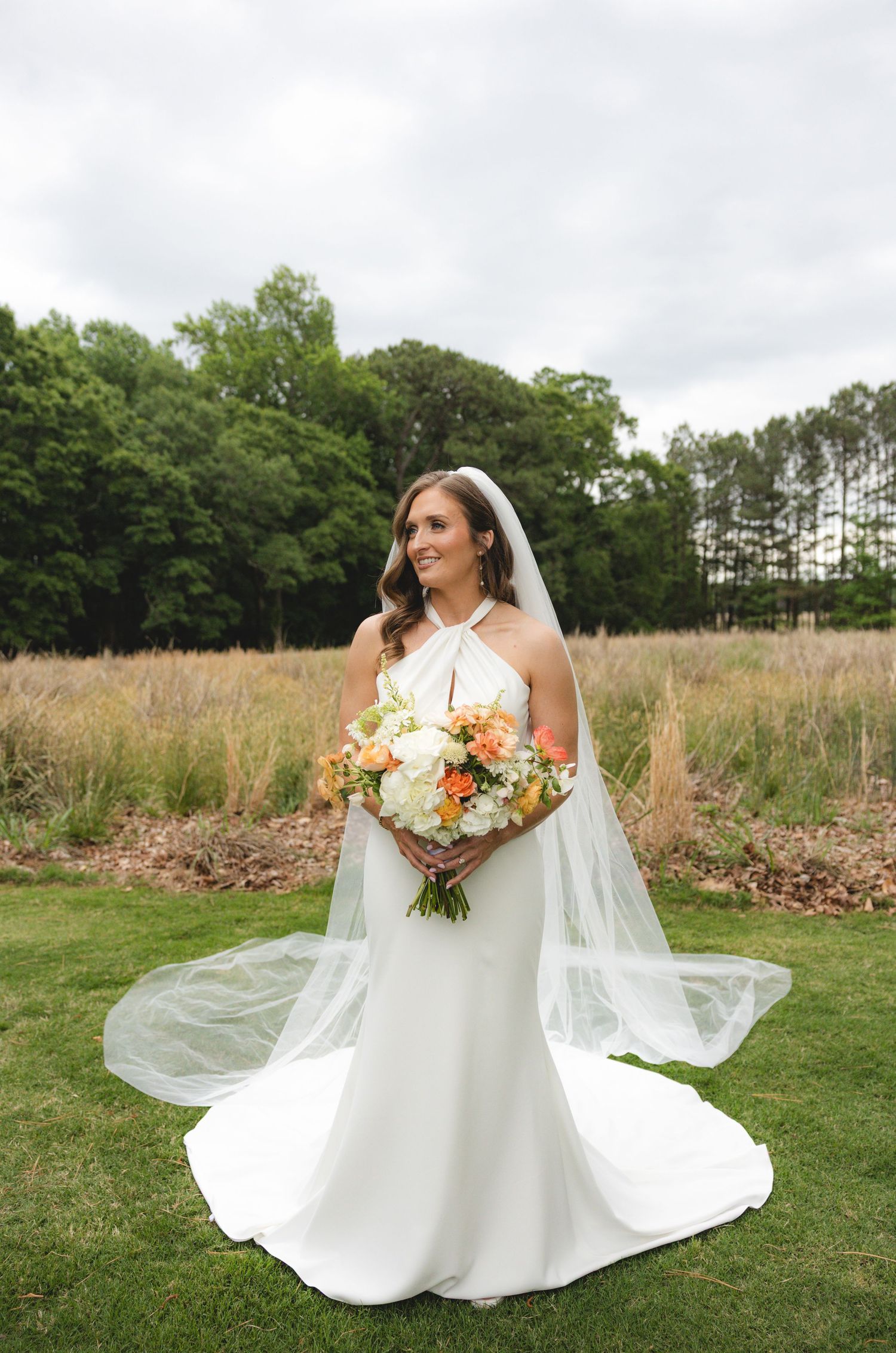 Bride in white halter wedding gown with long veil holds peach and white bouquet while smiling in a garden.